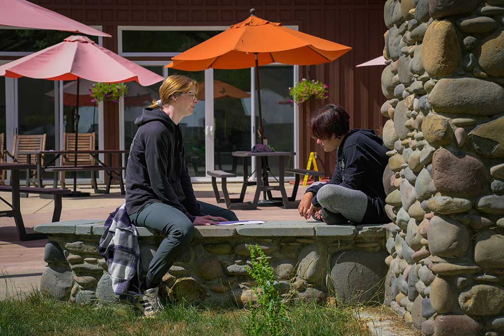 A woman sitting on the patio of the dining hall, talking with a camper during Compass camp