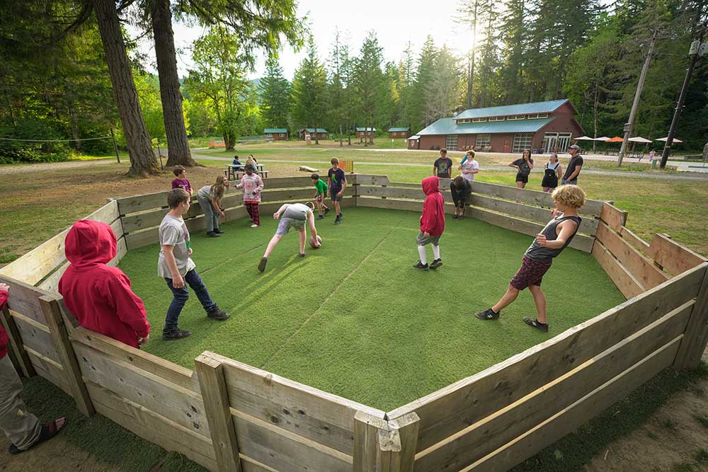 Campers playing gaga ball with the back field, dining hall, and upper cabins in the background